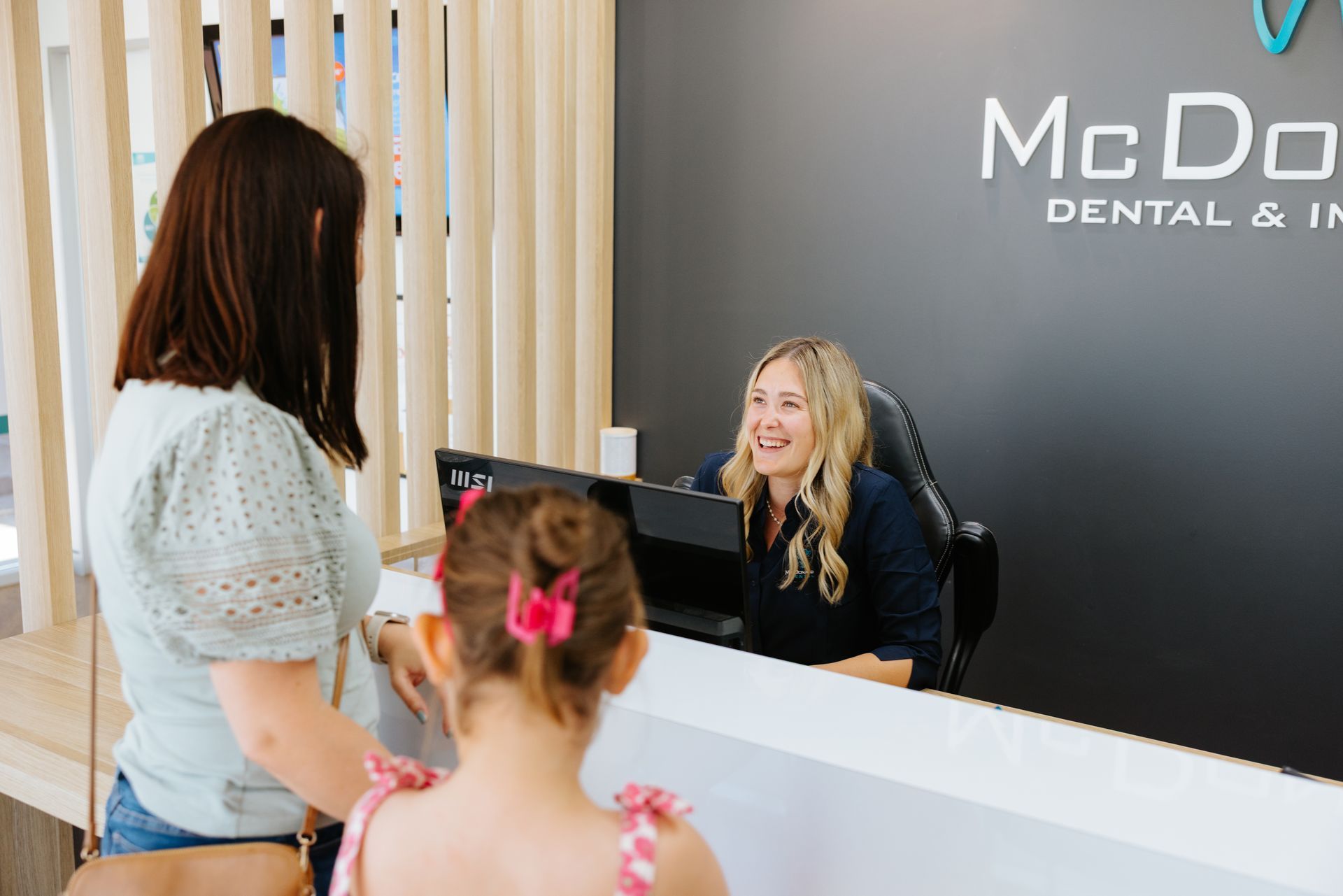 Woman and child at dental office reception desk. Receptionist smiles at them. — McDonald Dental in Gympie, QLD