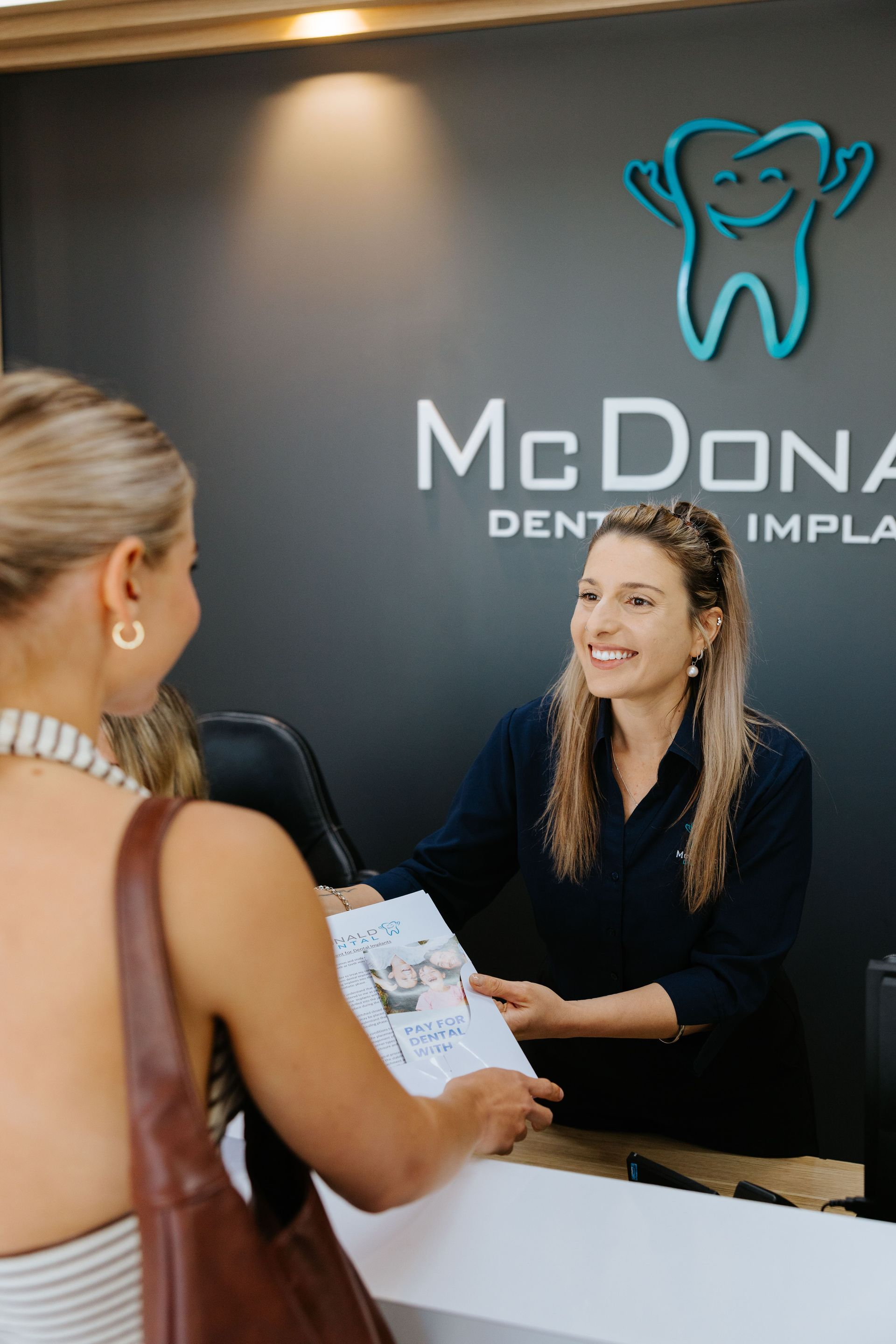 Woman at dental office desk hands brochure to a patient. Office logo visible. — McDonald Dental in Gympie, QLD