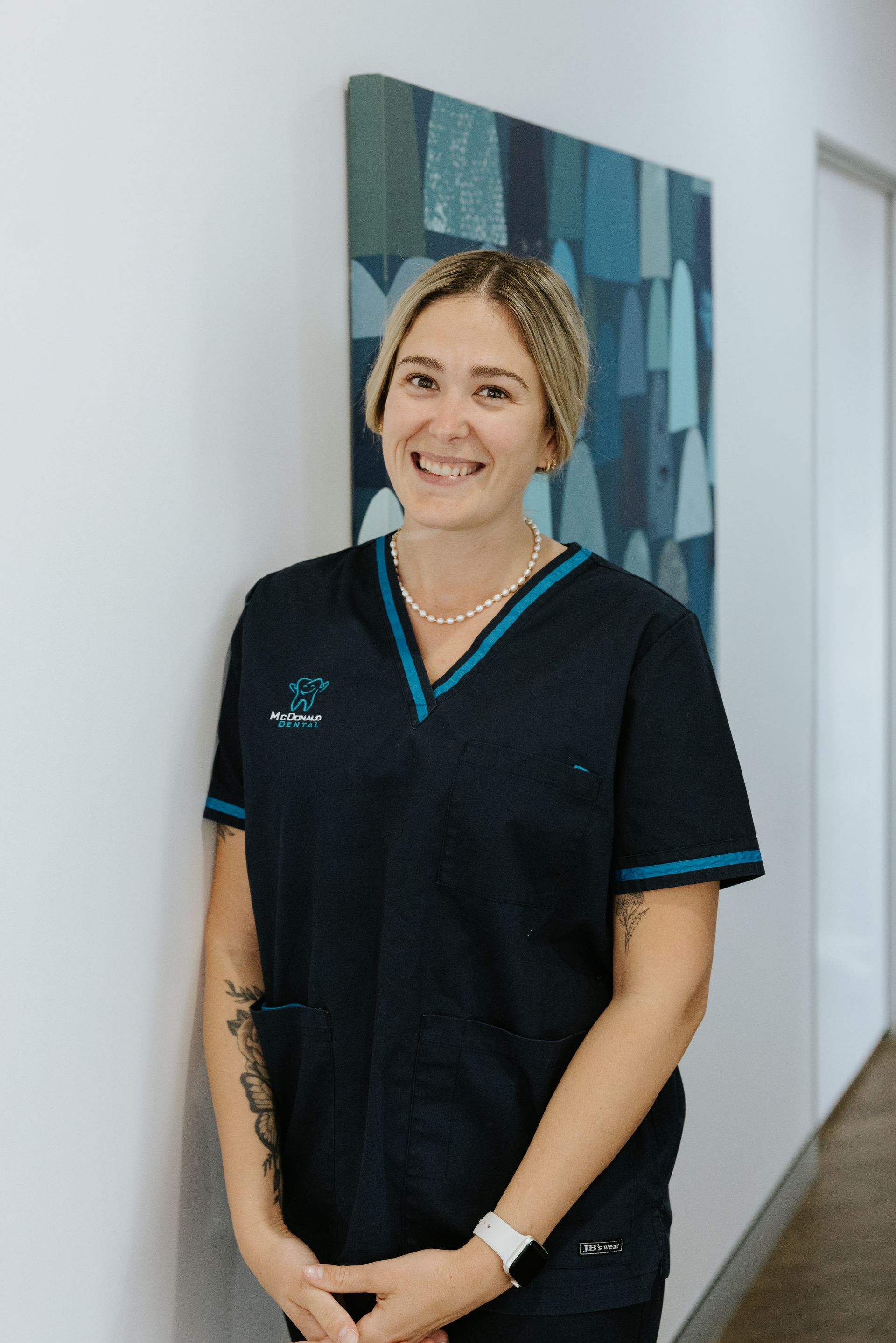 Woman in gray medical scrubs, hands in pockets, smiling. Light blue trim and logo on top. — McDonald Dental in Gympie, QLD