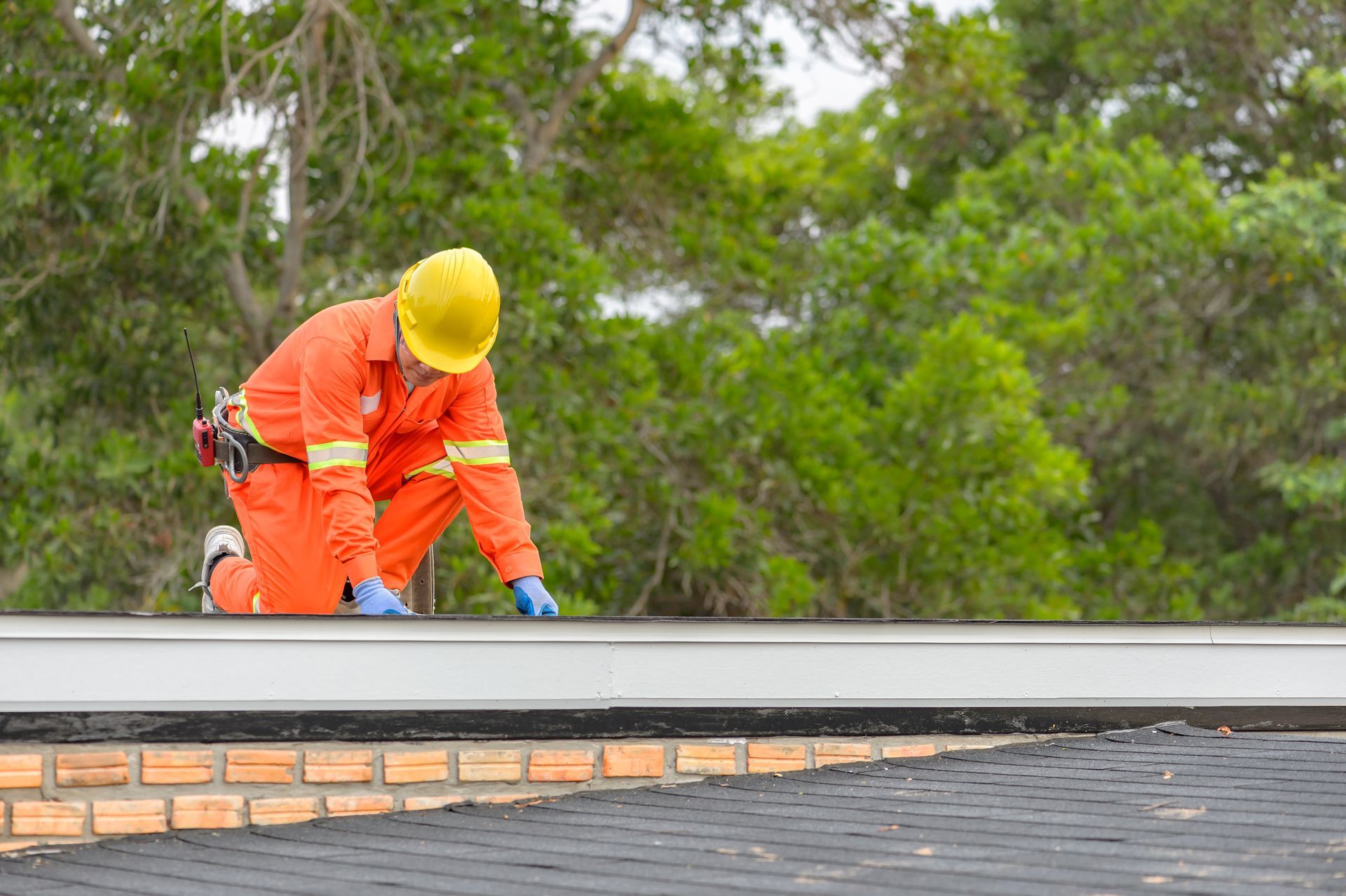 Worker in orange safety gear kneeling on roof edge during outdoor maintenance.