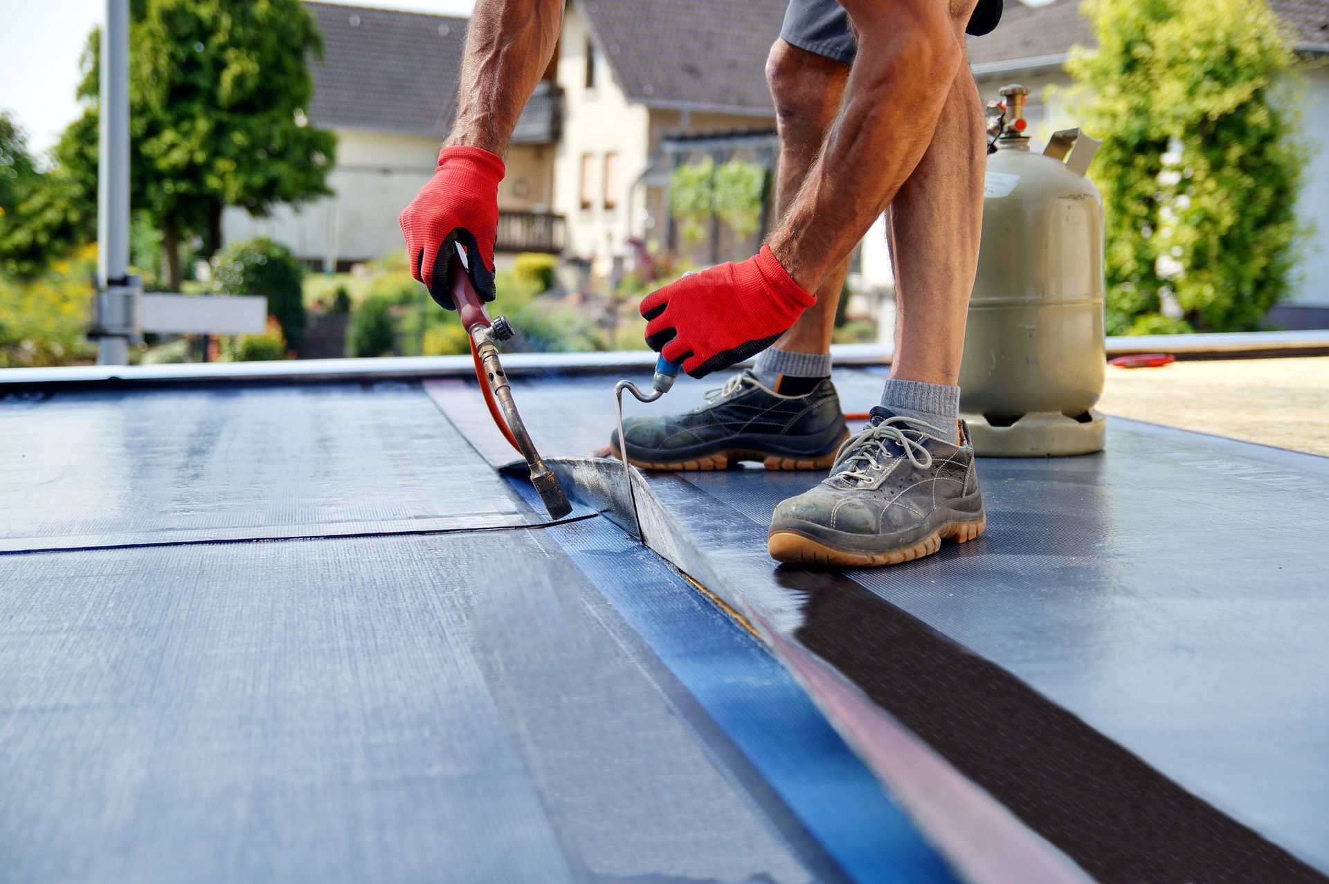 Roofer applying sealant to a flat roof, wearing red gloves and safety shoes, with a propane tank nearby.
