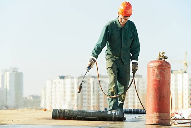 Roofer in green safety gear applies roofing felt with heat to a flat roof.