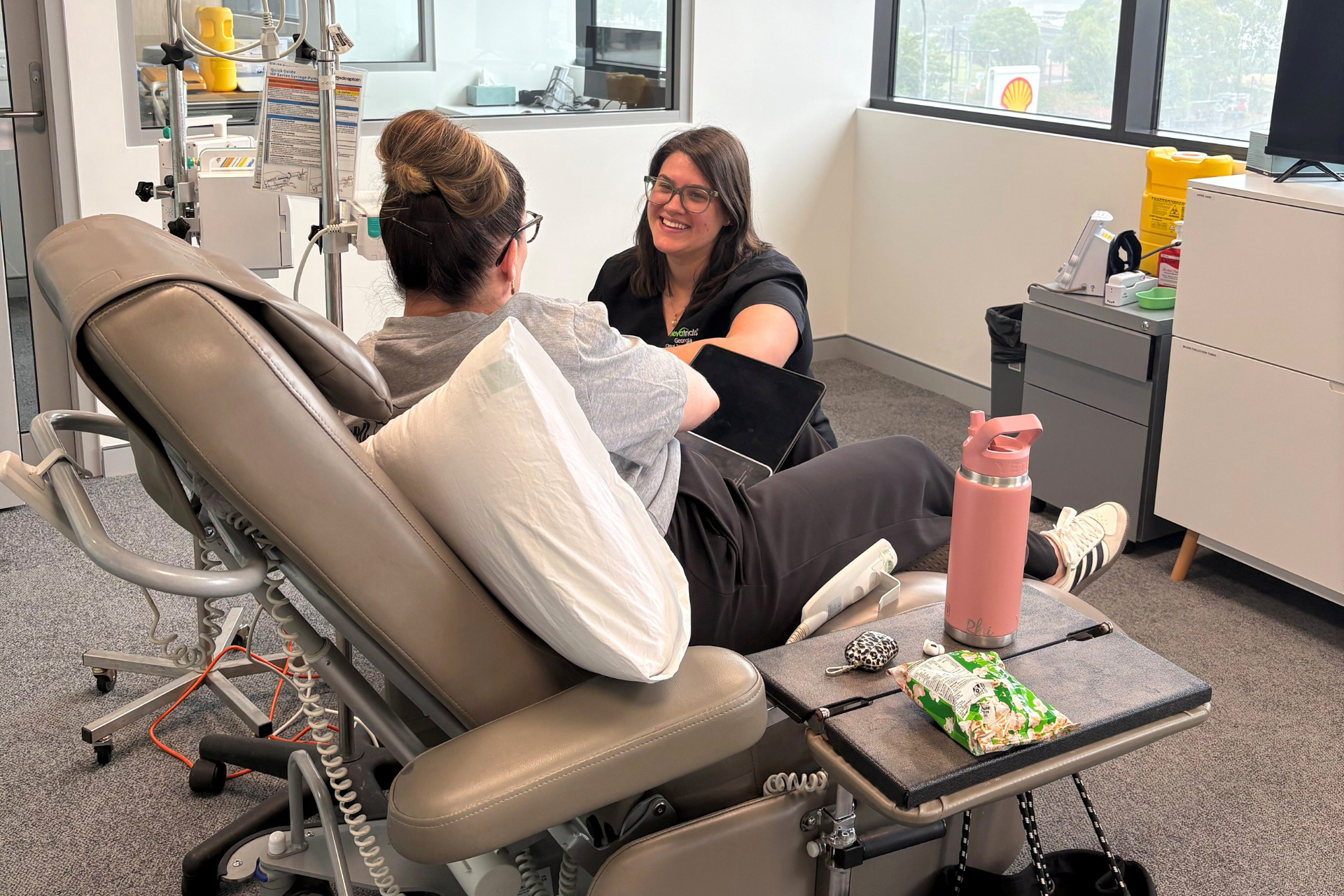 Person in medical chair talking with a healthcare worker. Room setting, patient is wearing glasses.