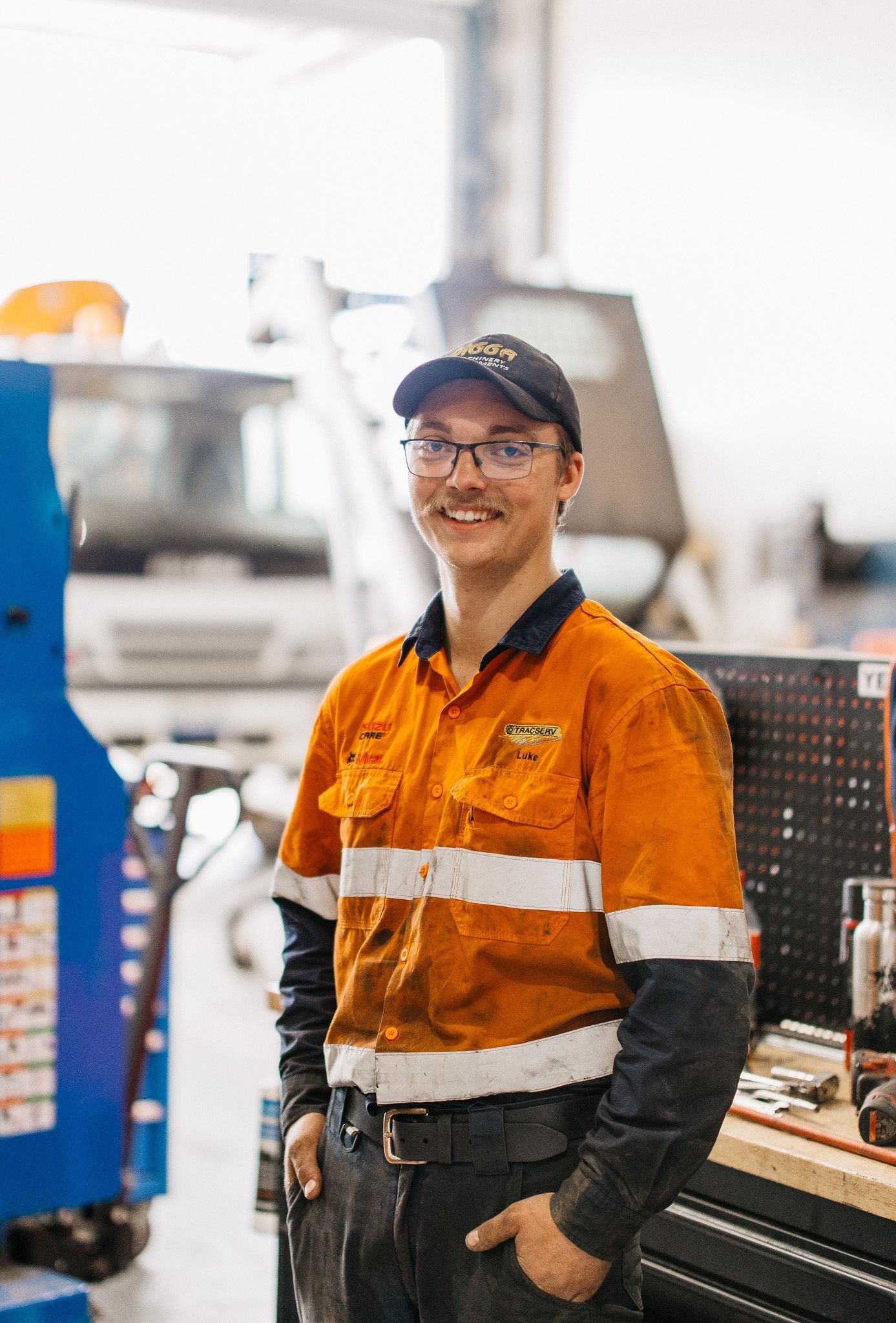 Mechanic in orange high-vis shirt smiles in a workshop, hands in pockets, wearing a cap.
