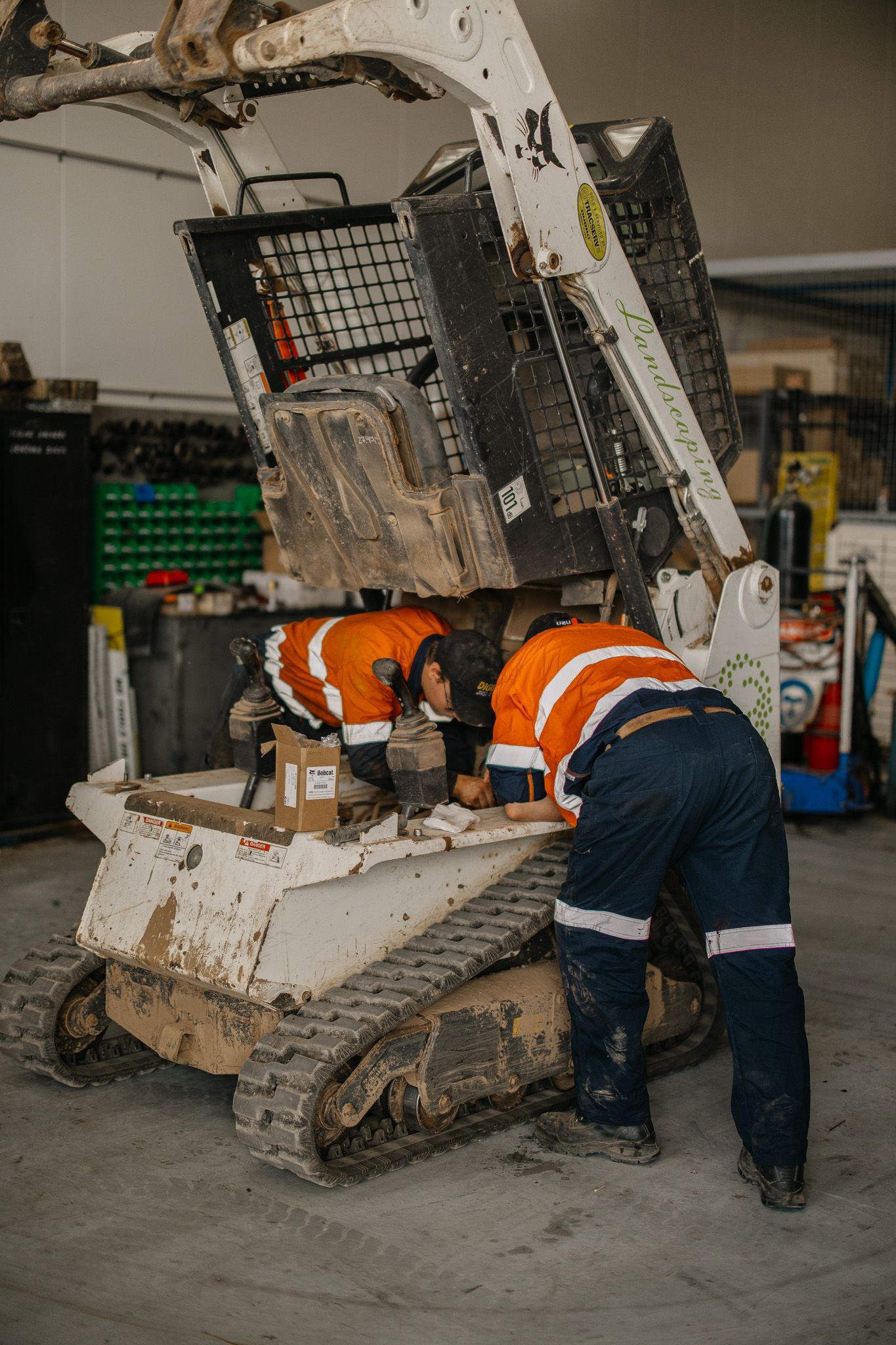 Two workers in orange safety vests and blue pants work on a Bobcat skid-steer loader in a garage.