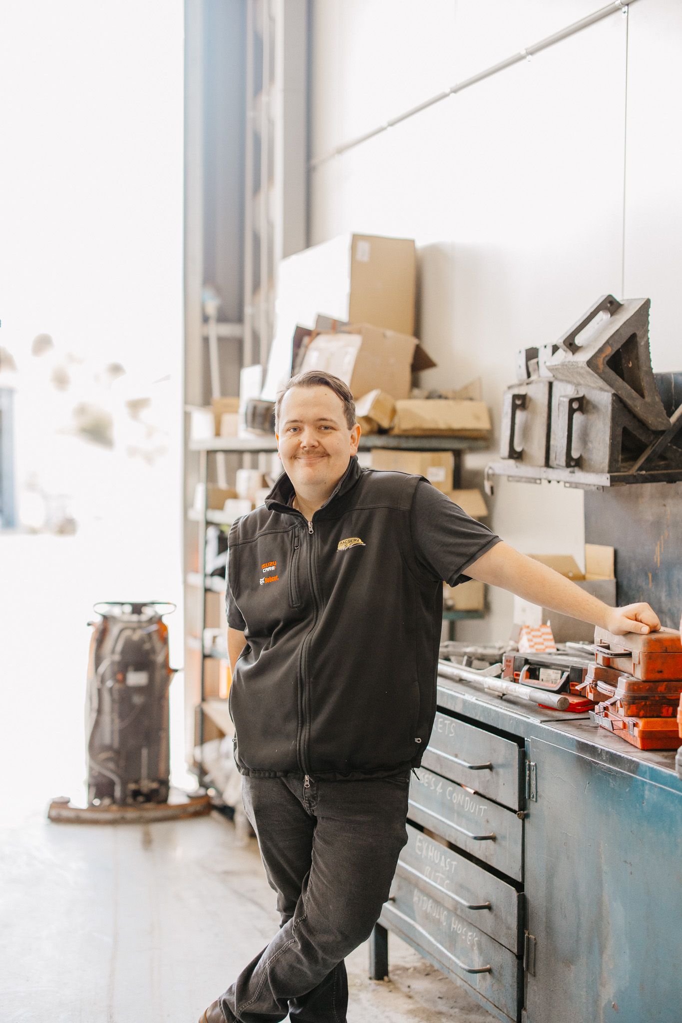 Man leaning against a work table in a workshop, smiling. He wears a black vest and jeans.
