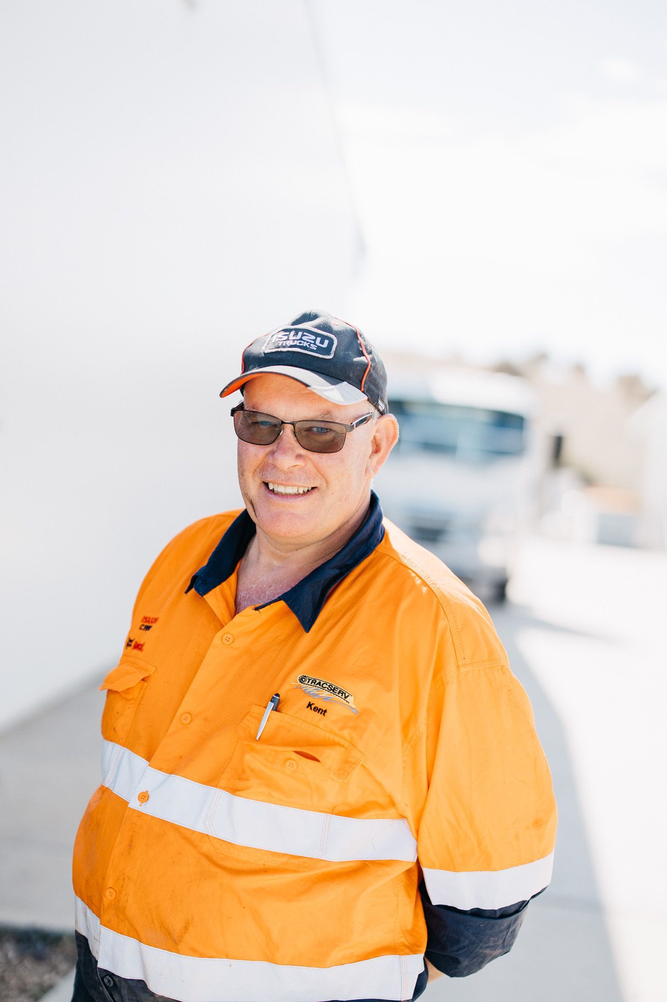 Man in orange work shirt and cap smiling outdoors with a vehicle in the background.