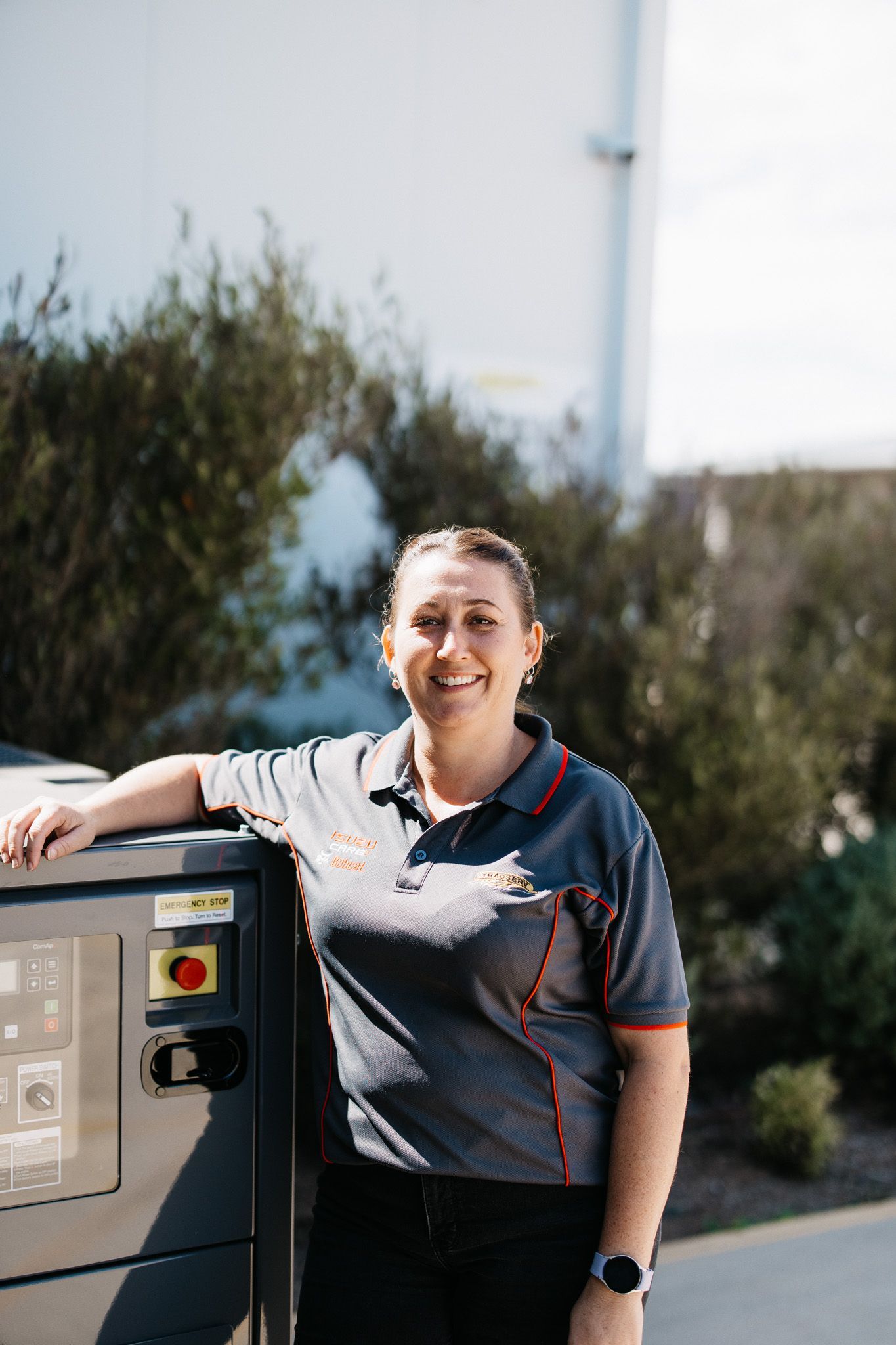 Woman in a grey uniform smiles, leaning on machinery outdoors near building and greenery.