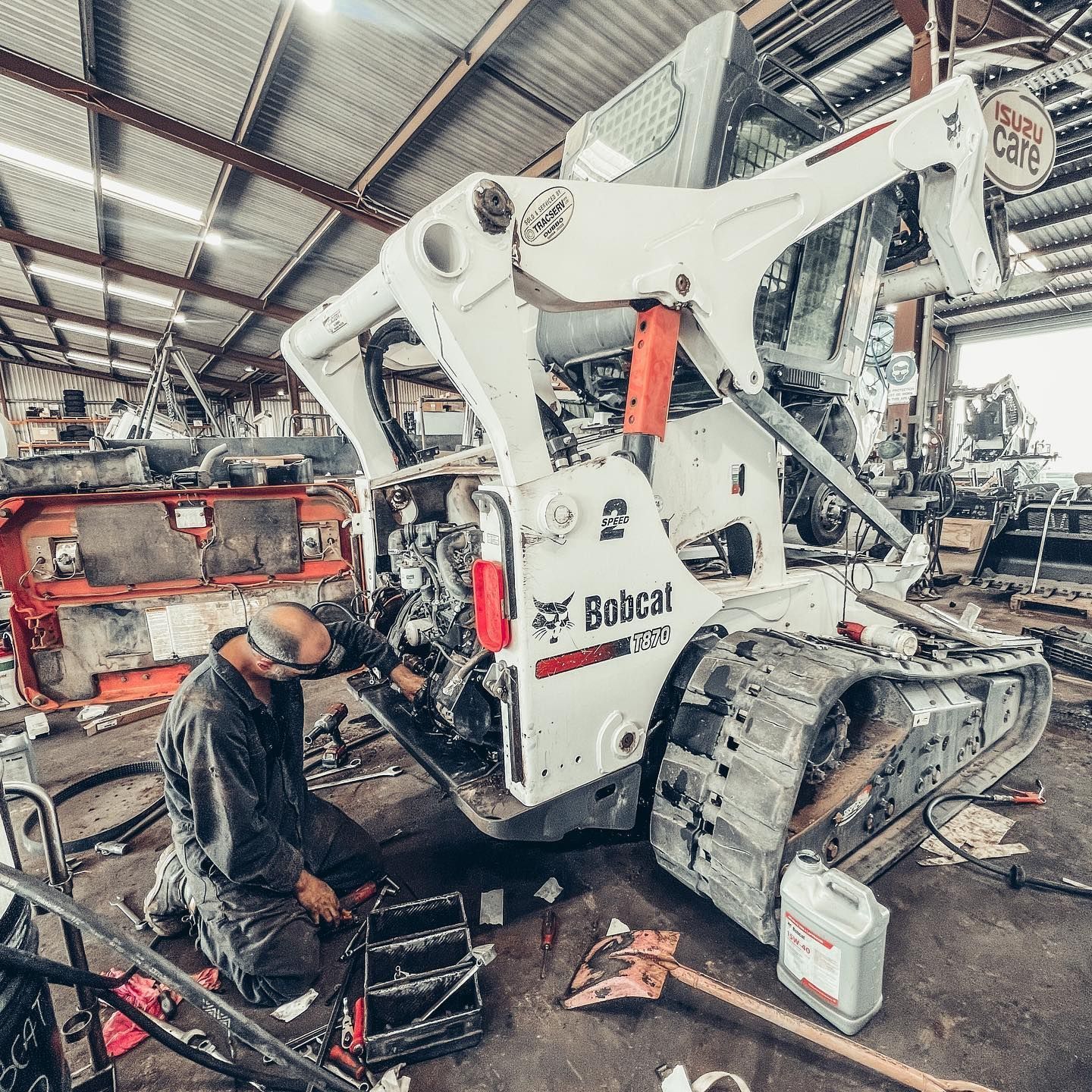Mechanic working on a white Bobcat skid steer in a cluttered garage, with tools and parts visible.