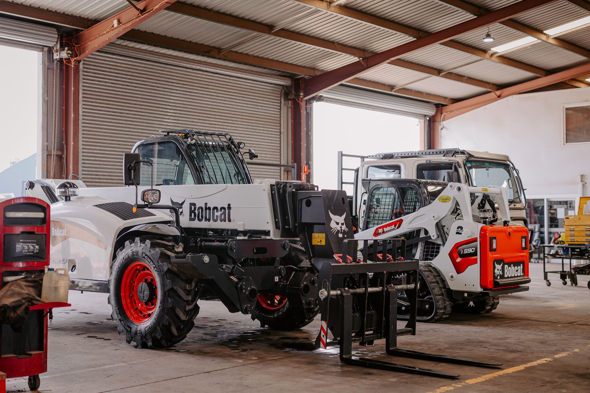 A bobcat tractor is parked in a garage next to a forklift.