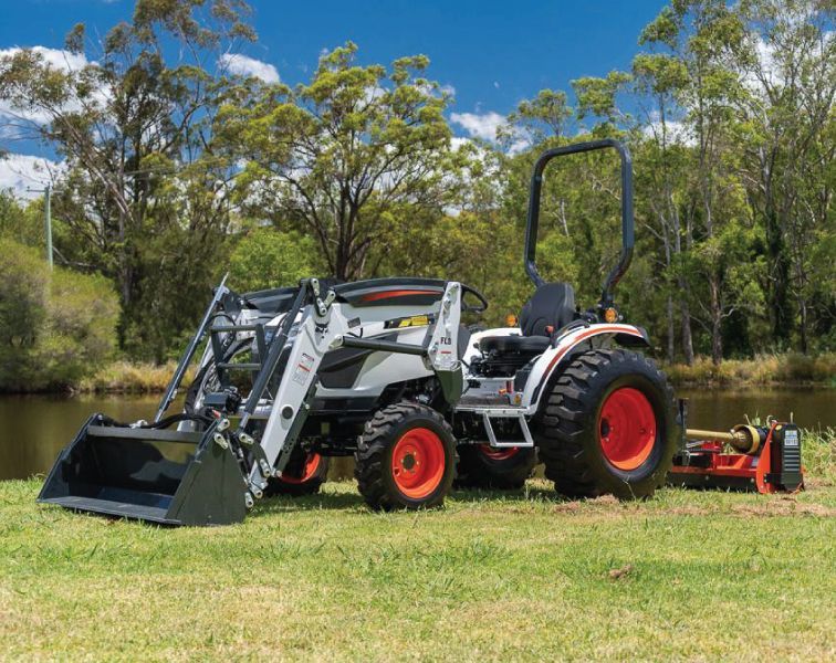 Compact tractor with front loader and rear rotary cutter parked on grass near water.