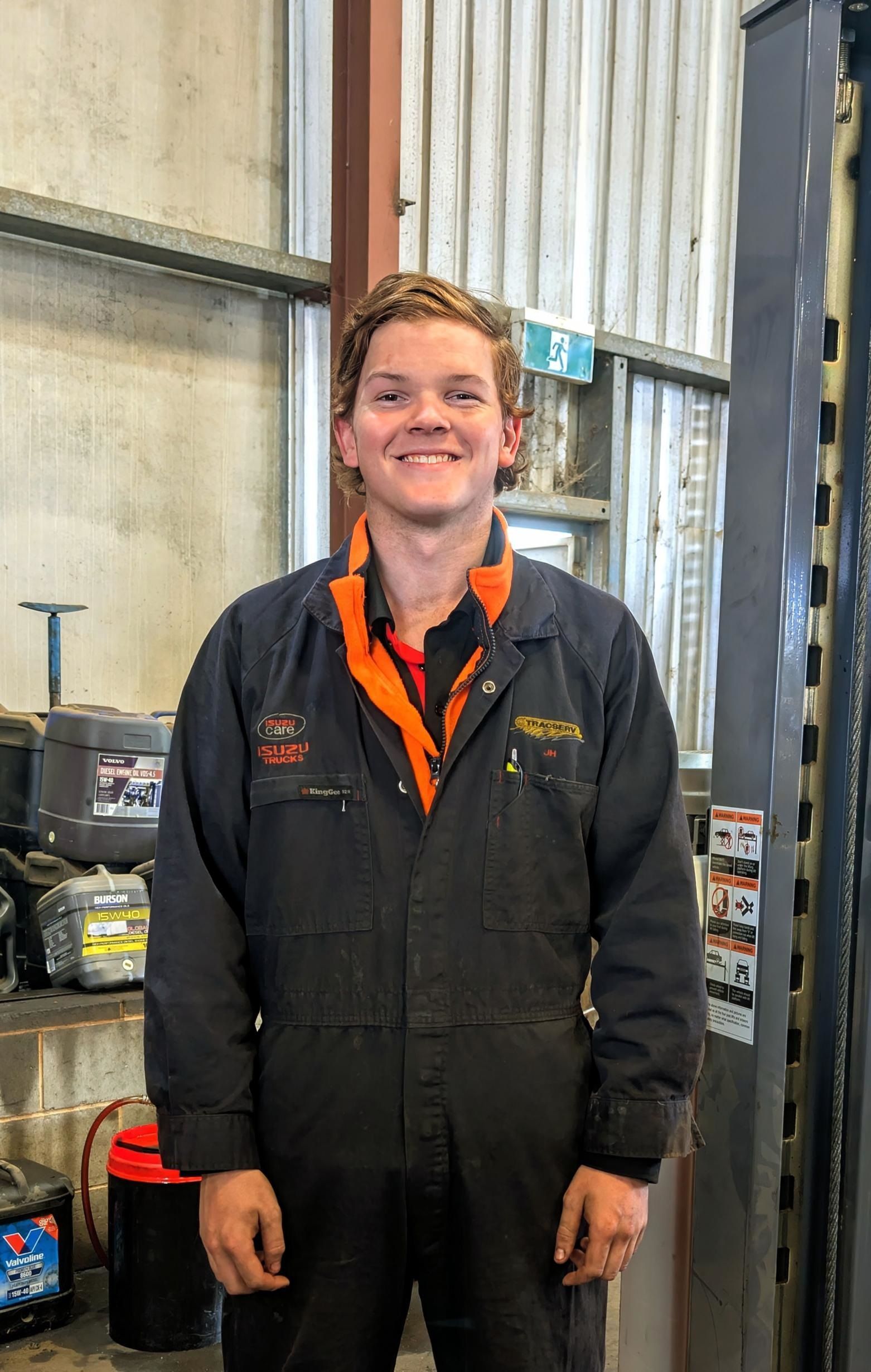 Mechanic in blue jumpsuit, orange trim, smiling in a garage setting with equipment.