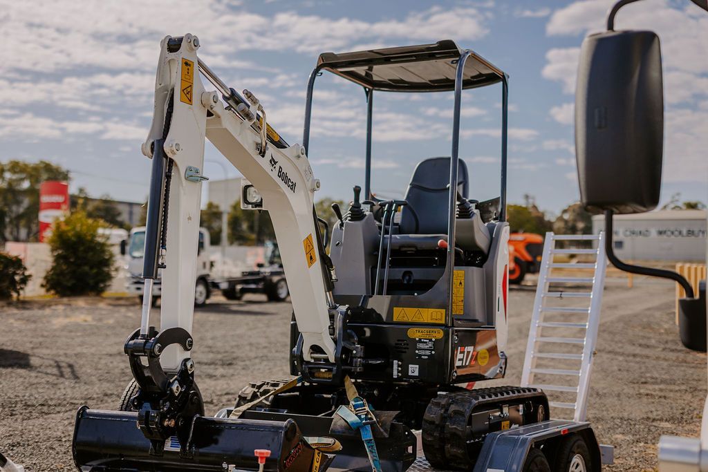 A Small Excavator Is Parked In A Gravel Lot Next To A Trailer — Tracserv in Dubbo, NSW