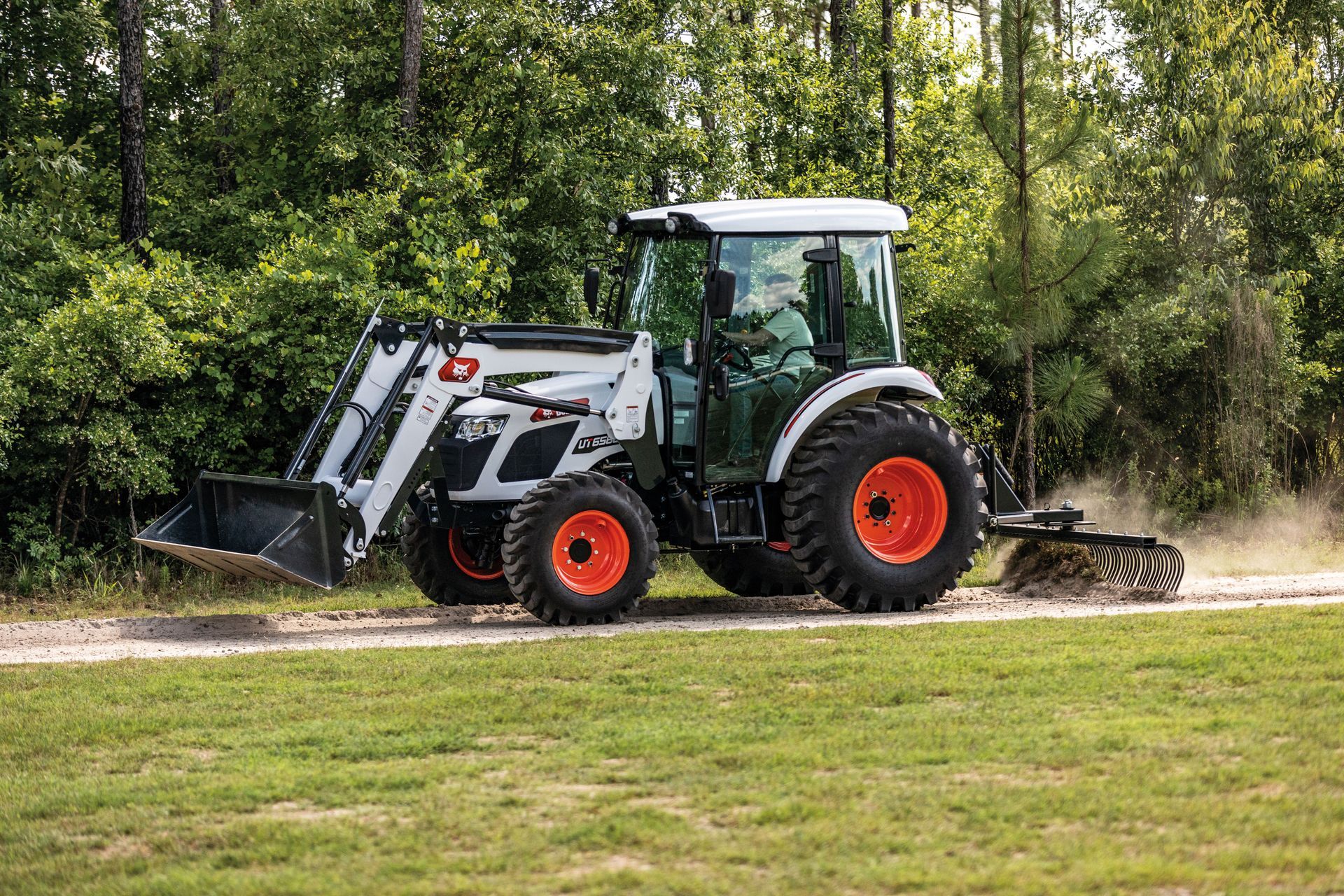 Bobcat tractor on a dirt road, raking up debris. White and orange, in a wooded area.