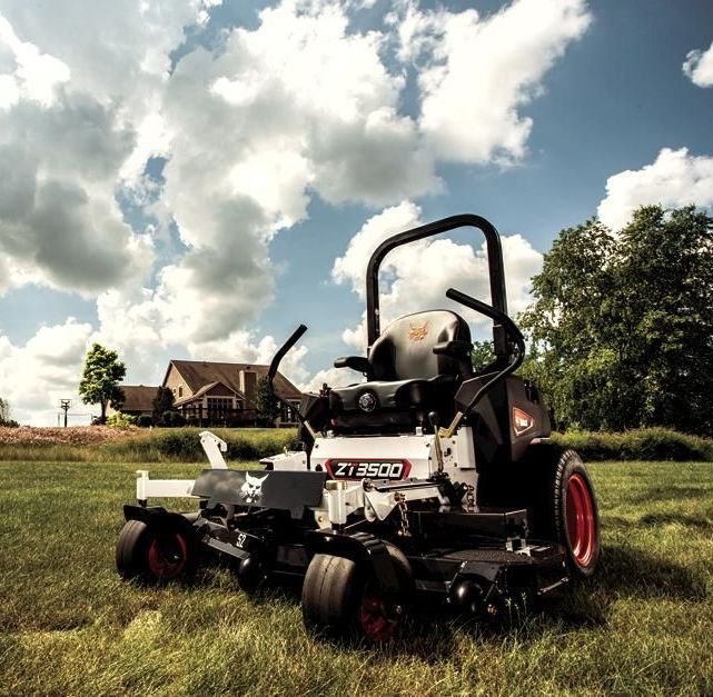 White and black zero-turn mower on green grass with house and blue sky in background.