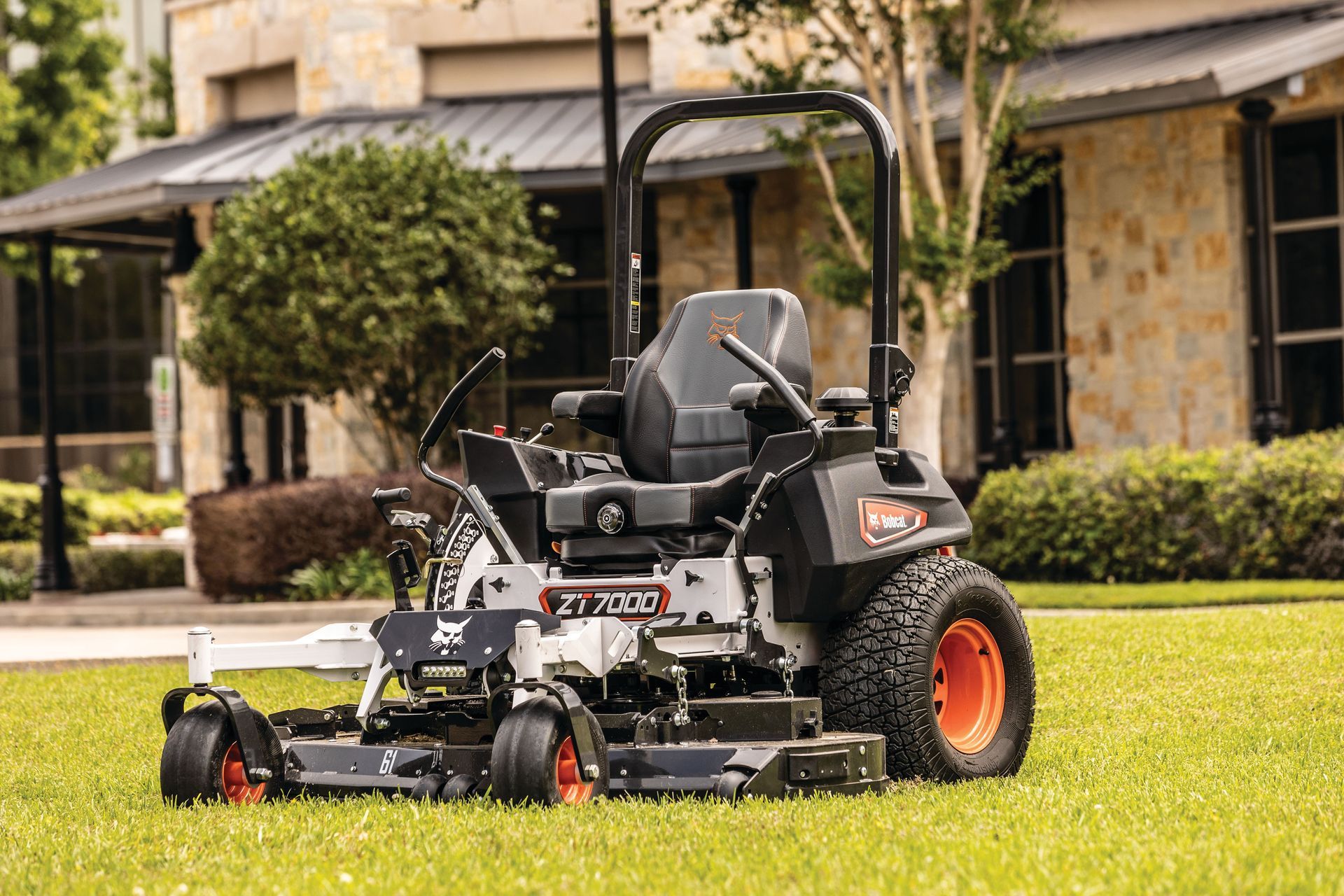 Bobcat zero-turn mower on green grass; orange wheels, black frame, in front of a building with stone siding.