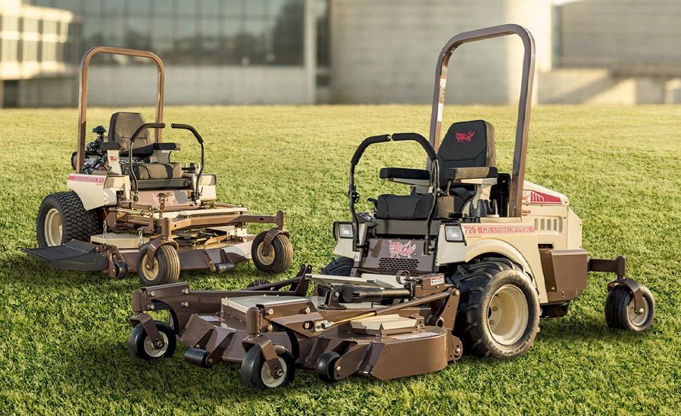 Two brown and cream-colored zero-turn lawn mowers on a grassy field; in front of a building.