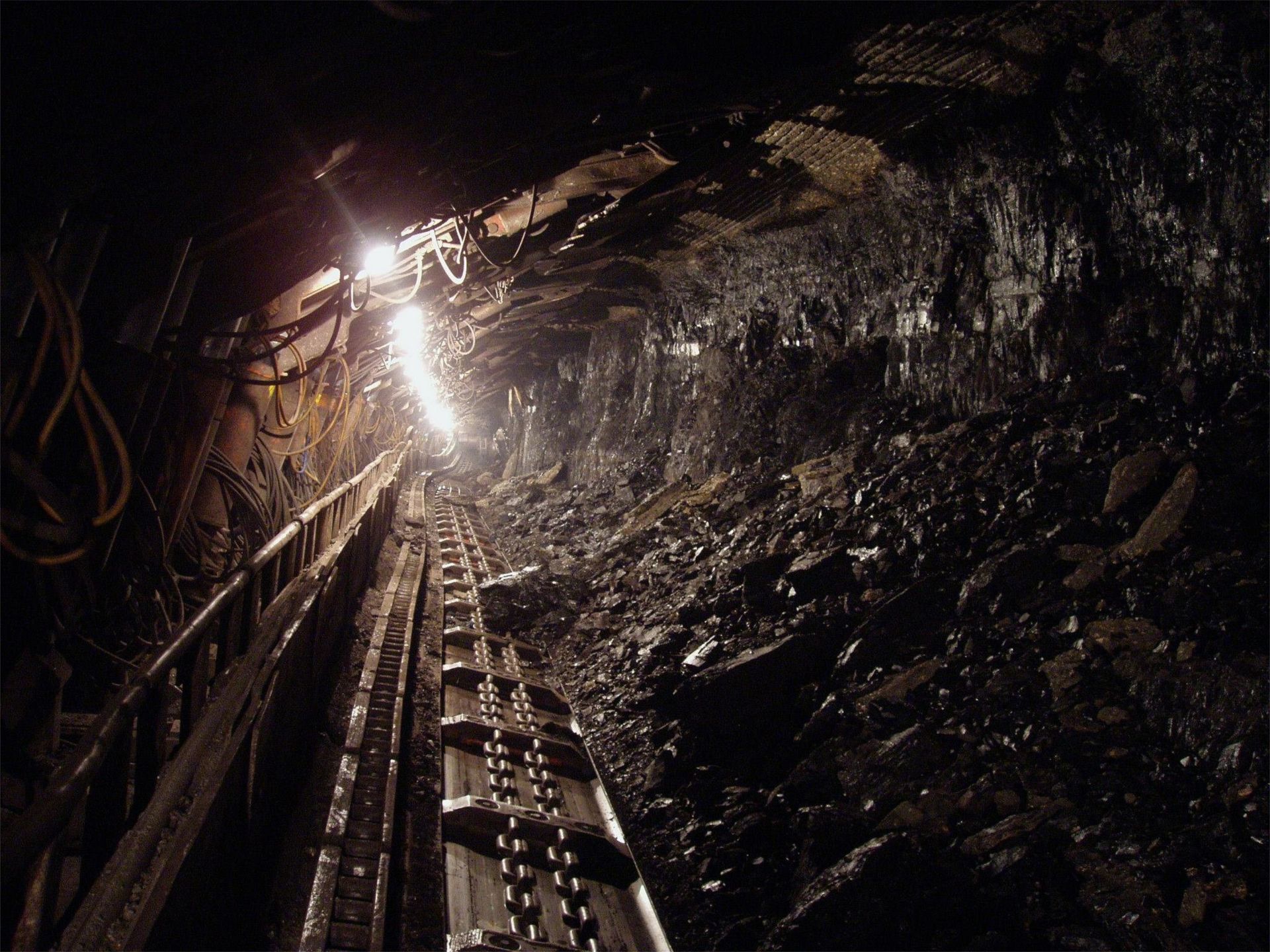 Inside a dark coal mine tunnel with tracks and coal debris illuminated by bright lights.