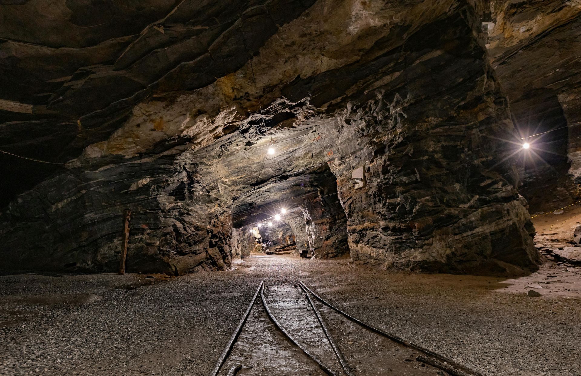 Dark mine tunnel with train tracks. Arched opening with lights, leading to another section of the mine.