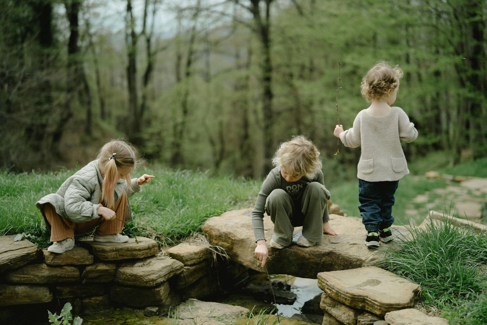 Three children exploring a small pond in a wooded area, squatting and looking at the water.