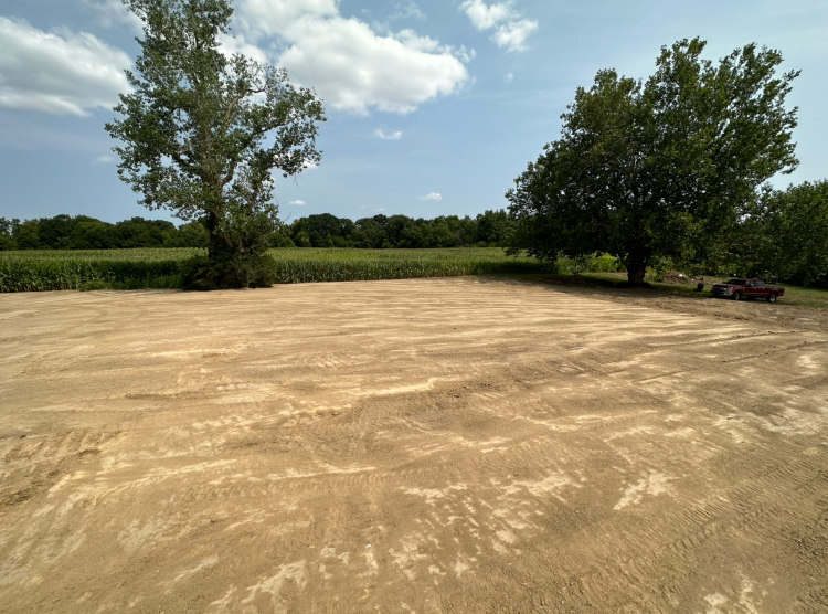 Open, sandy lot with two trees and a cornfield in the background under a partly cloudy sky.