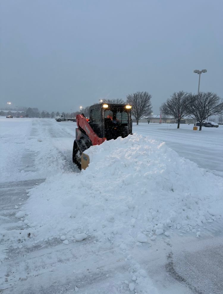 Snowplow clearing a parking lot during a snowstorm. The plow pushes a large pile of snow, with bare trees and streetlights in the background.