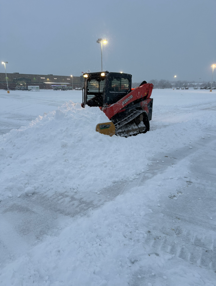 Red skid steer plowing snow in a parking lot on a snowy day. Black cab, yellow blade, and tracks visible.