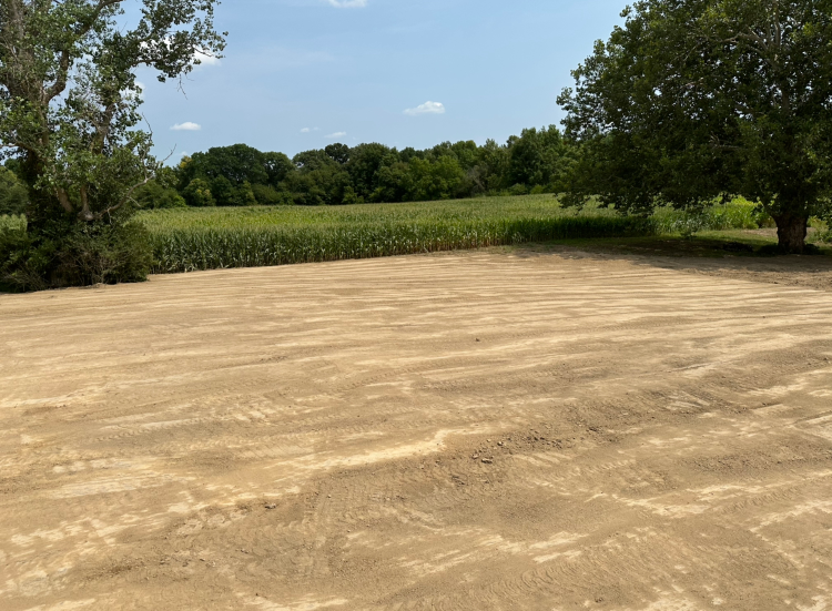 A cleared, sandy area with a field of green crops and trees in the background under a blue sky.