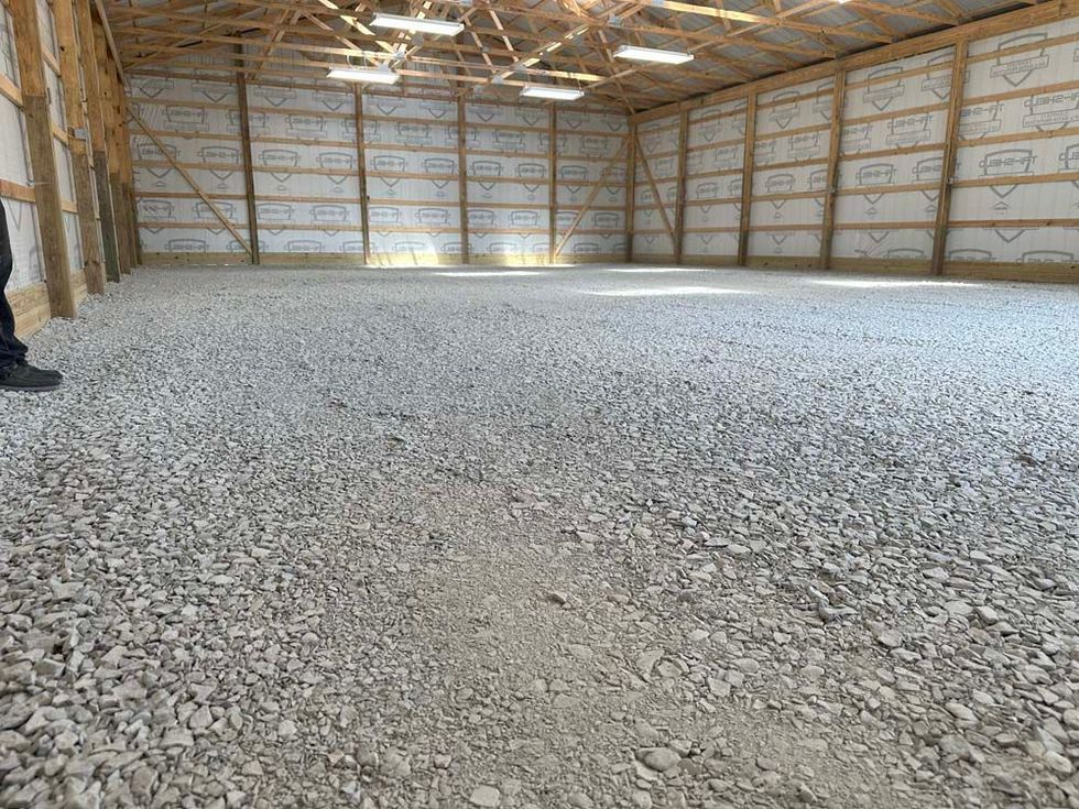 A large, empty barn interior filled with gravel flooring. Wooden beams frame the walls and ceiling.