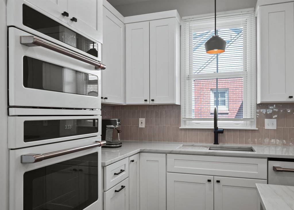 A kitchen with white cabinets , a double oven , a sink , and a window.