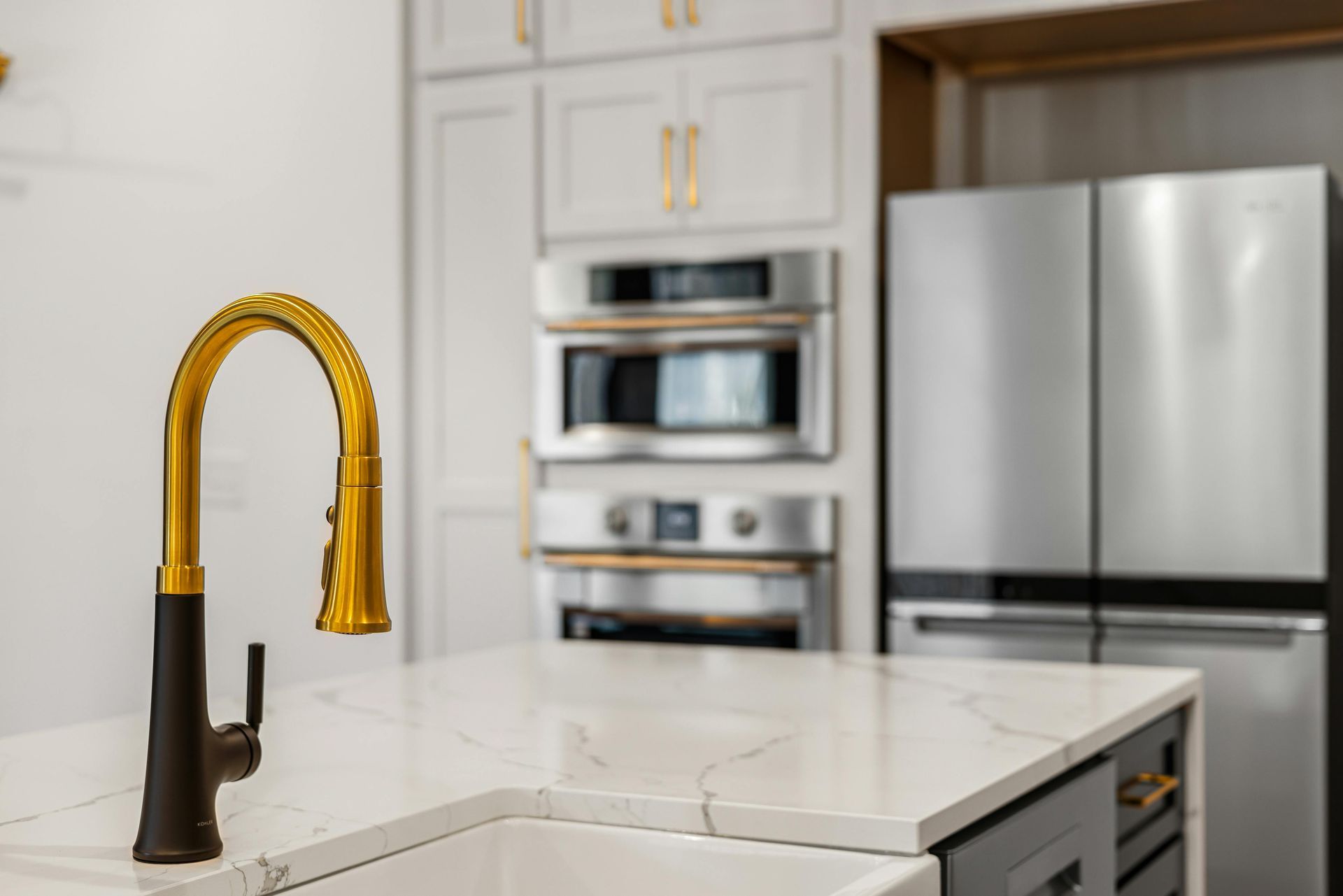 A kitchen sink with a gold faucet and a stainless steel refrigerator in the background.