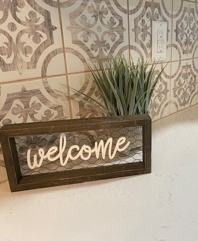 A welcome sign is sitting on a counter next to a plant.