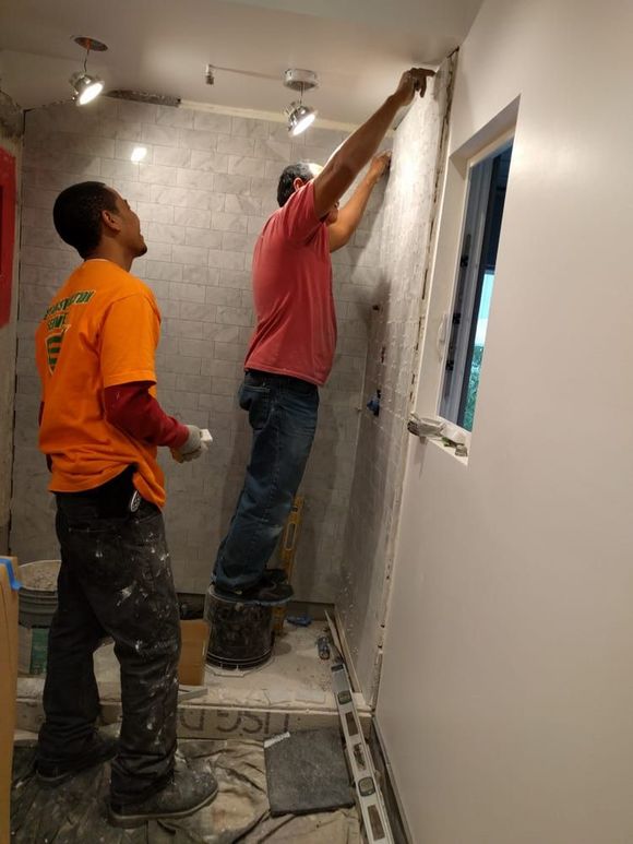Two workers tiling a bathroom. One on a bucket, reaching up. Other worker observing.