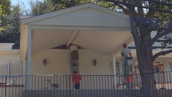 Workers painting a covered porch; one on a ladder, others using rollers. Exterior of a building with a tree.