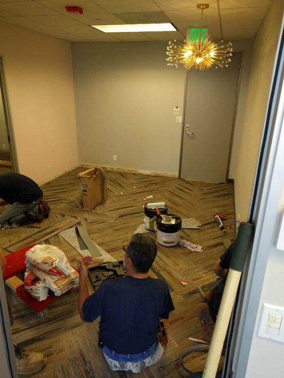 Workers installing flooring in a room with pink and gray walls, tools, and a decorative light fixture.