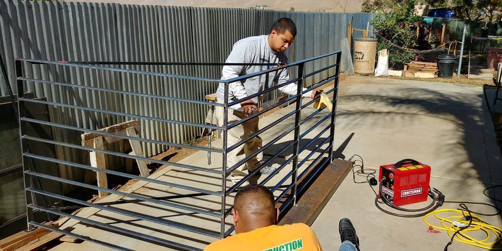 Two men are working on a metal railing, one welding and another seated. A red welding machine is on the ground.