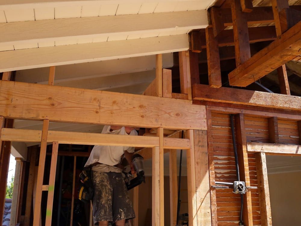 Construction worker nailing lumber in a partially framed structure with exposed beams and electrical components.