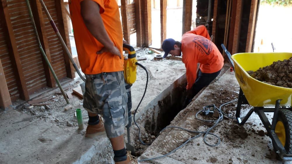 Two workers in orange shirts excavate indoors; one drills, one shovels dirt, near a wheelbarrow.