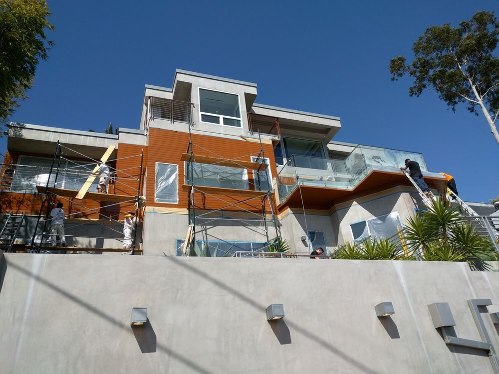 House under construction; scaffolding and workers visible. Orange, white, and grey exterior; blue sky.