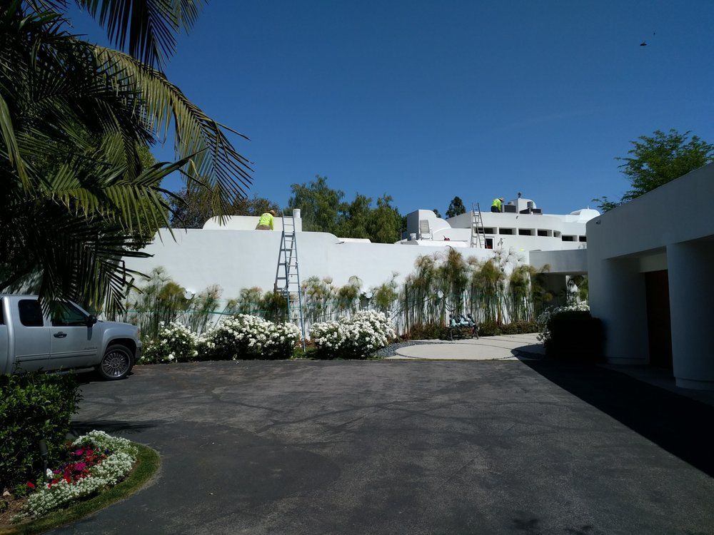 White building with workers on the roof, surrounded by foliage, a truck parked nearby, and a driveway.