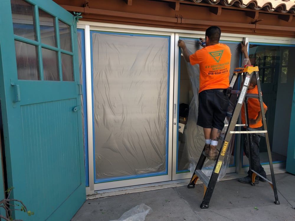 Two men installing plastic sheeting on a sliding glass door, using a ladder.