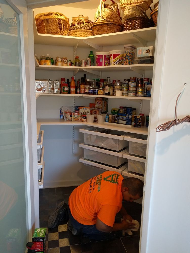 Person in orange shirt kneeling in pantry, adjusting flooring. Pantry has shelves, baskets, and food.