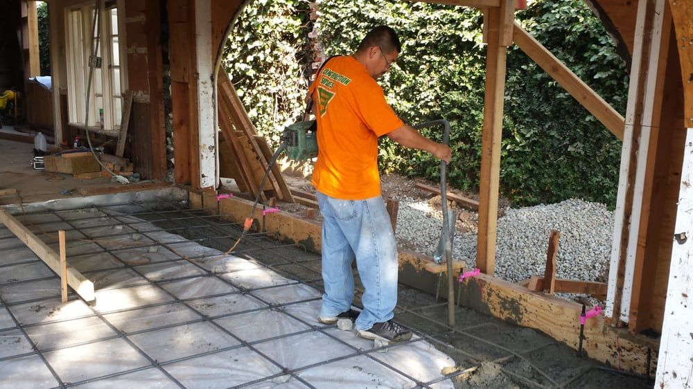 Construction worker in orange shirt smoothing wet concrete, inside building frame.