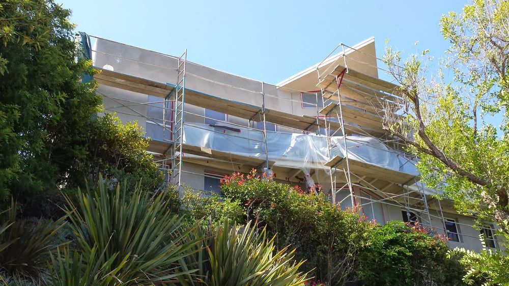 Building under construction with scaffolding and protective wrap. Green foliage surrounds.