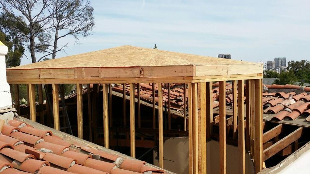 Framed wooden structure atop a terracotta-tiled roof, under construction, with a city skyline in the background.