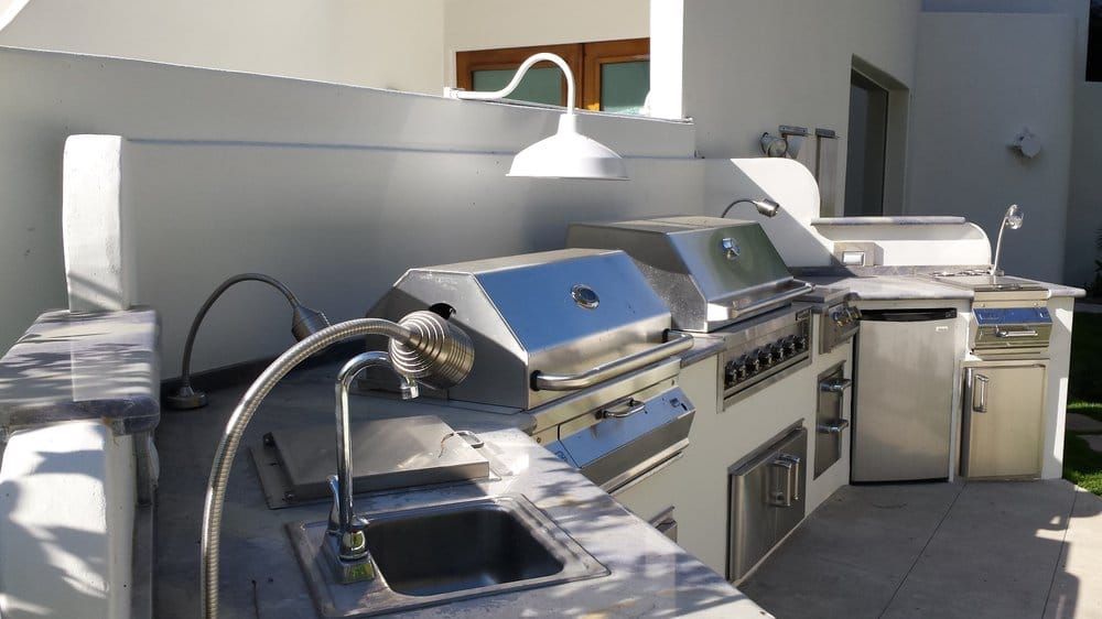 Outdoor kitchen with stainless steel grills, sink, and refrigerator, built into a white structure.