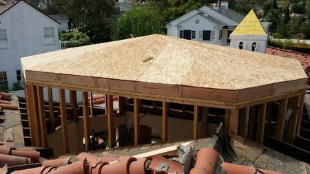 Framing of a light brown wooden structure under construction on a clay tile roof, with surrounding buildings.