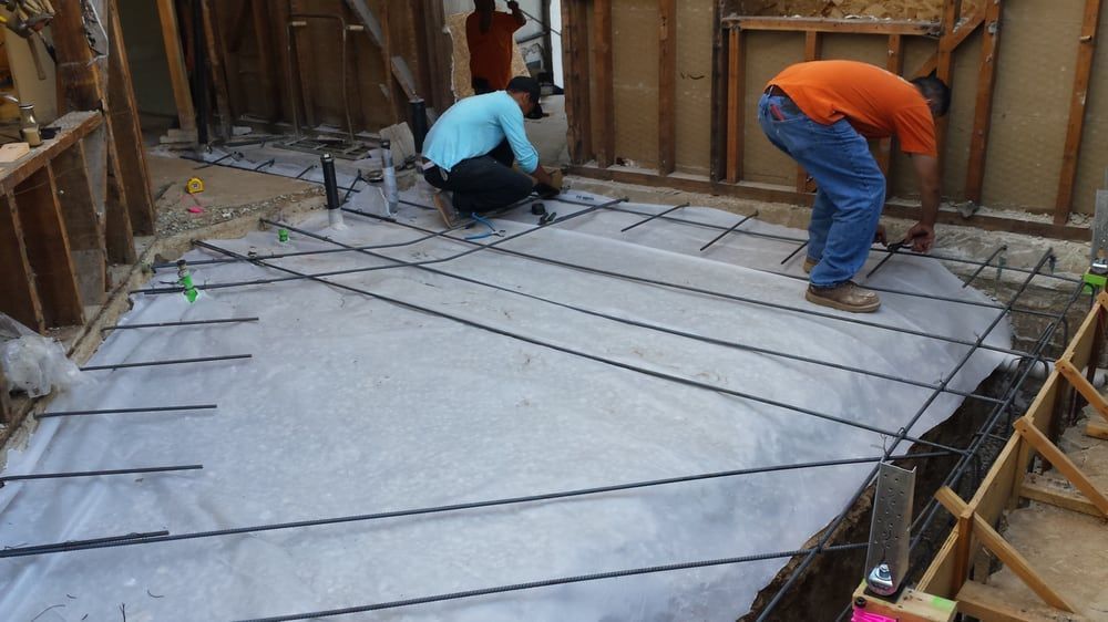 Construction workers placing rebar grid on a plastic sheet, preparing for concrete floor in a partially demolished room.