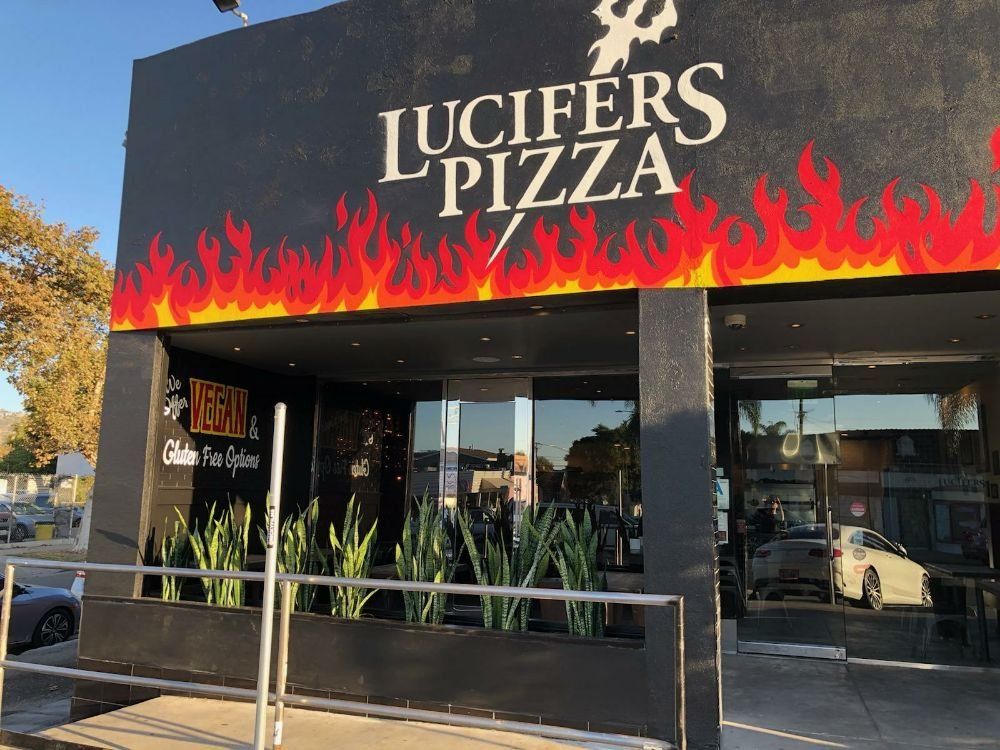 Lucifer's Pizza storefront with flames. Black exterior, white logo, glass windows, plants and parked cars.