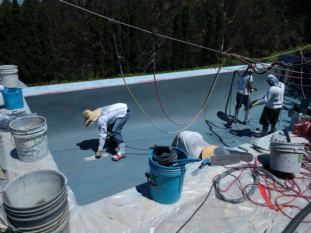 Workers applying a blue coating to a roof. Buckets, hoses, and protective gear are visible. Outdoors, sunny day.